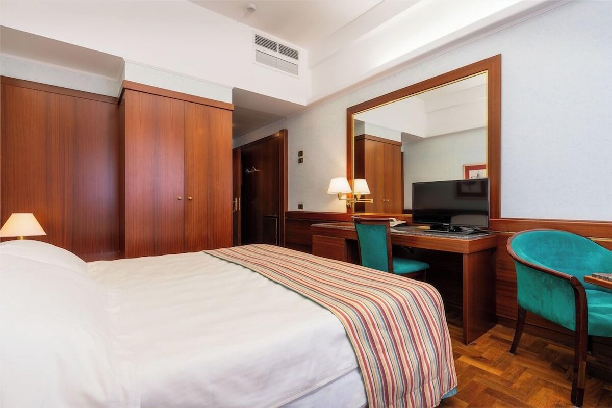 A hotel room with a bed, a desk with a computer, a chair, and a large mirror.