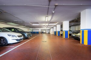 Un estacionamiento con varios coches aparcados en filas, pilares blancos con franjas azules e iluminación fluorescente en el techo.