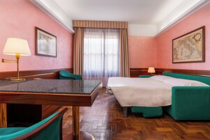 A hotel room with a wooden floor, a bed with green upholstered headboard, a desk with a lamp, and two chairs.