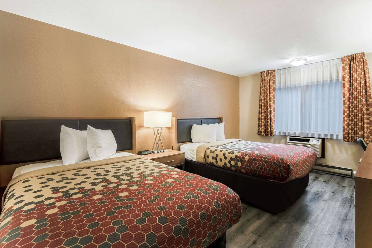 Ein Hotelzimmer mit zwei Betten, einem Nachttisch mit Lampe, einem Fenster mit Vorhängen und einer Klimaanlage.