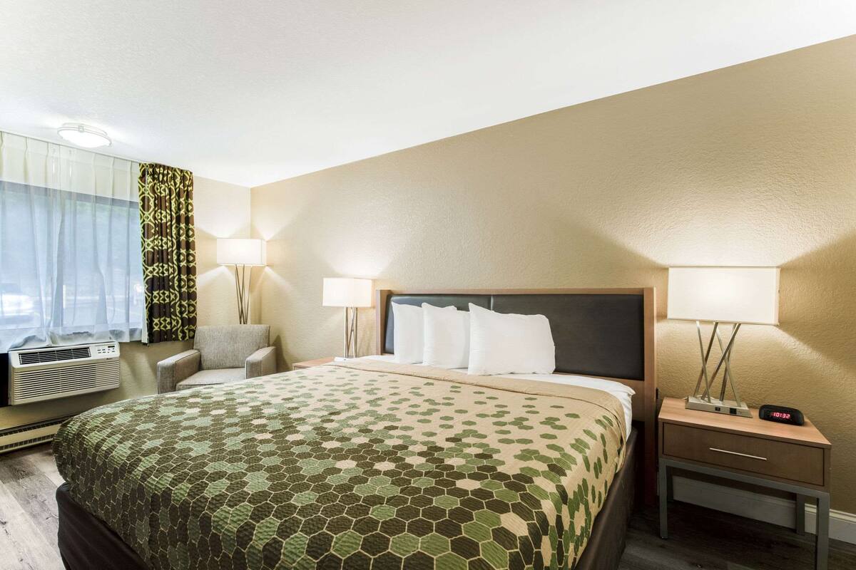 Ein Hotelzimmer mit einem großen Bett, zwei Nachttischlampen, einem Sessel, einem Schreibtisch und einem Fenster mit transparenten Vorhängen.