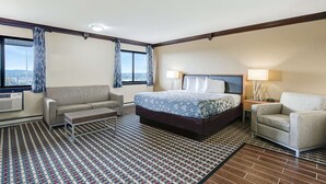 Habitación de hotel con una cama grande, un sofá, una mesita, una mesita de noche con lámpara y una ventana con cortinas.