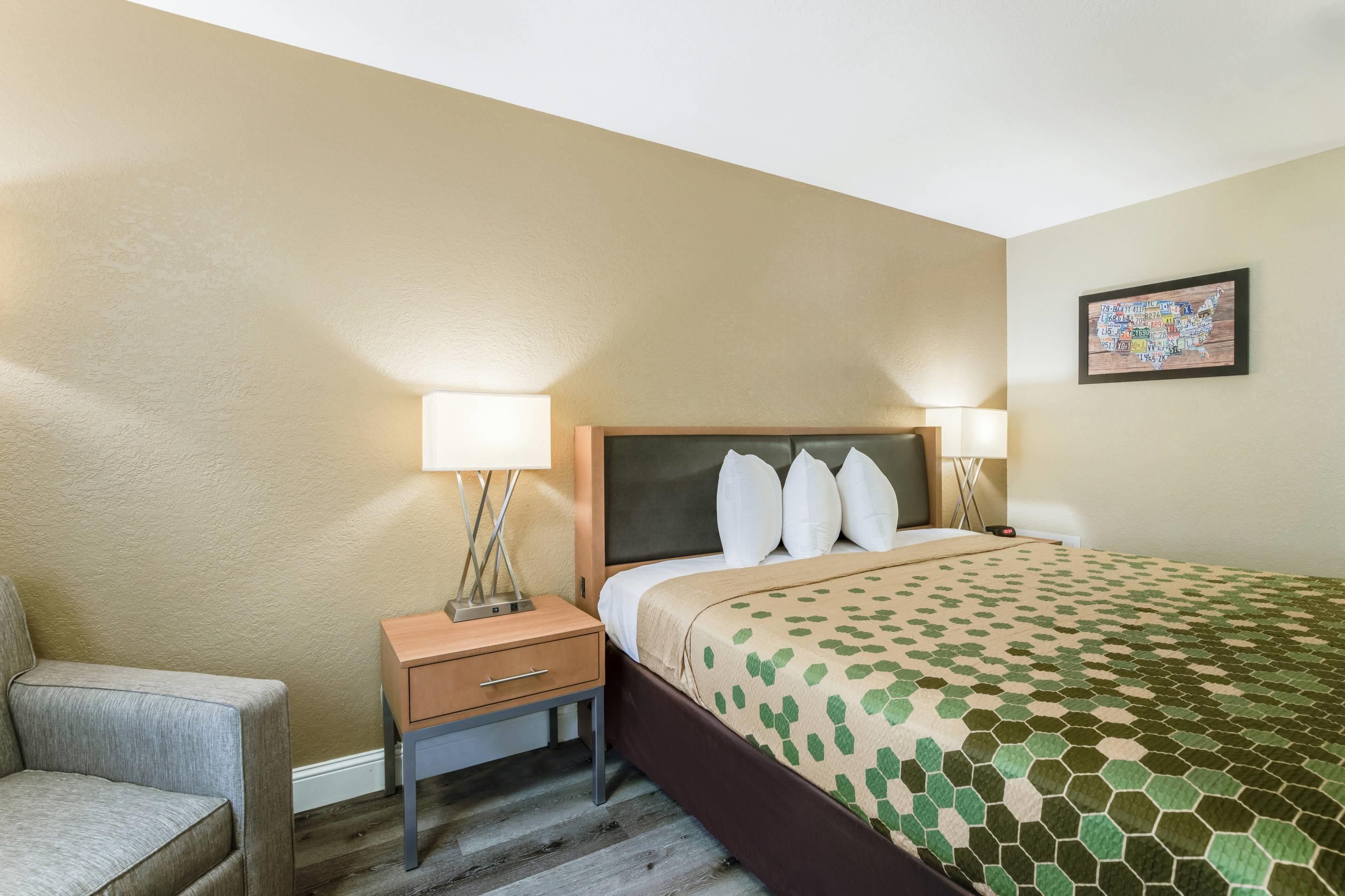 Quarto de hotel com cama, luminárias de cabeceira, mesa de cabeceira, uma cadeira e uma obra de arte na parede.
