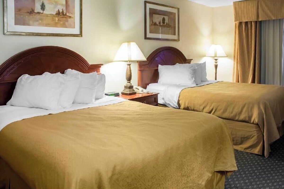 A hotel room with two beds, each with a wooden headboard, white bedding, and a lamp on the nightstand.