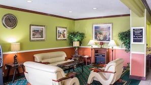 Vestíbulo de hotel con pared verde, un reloj, obras de arte enmarcadas, un sofá, dos sillones, una mesa de centro y un cartel que detalla servicios como desayuno de cortesía y gimnasio.