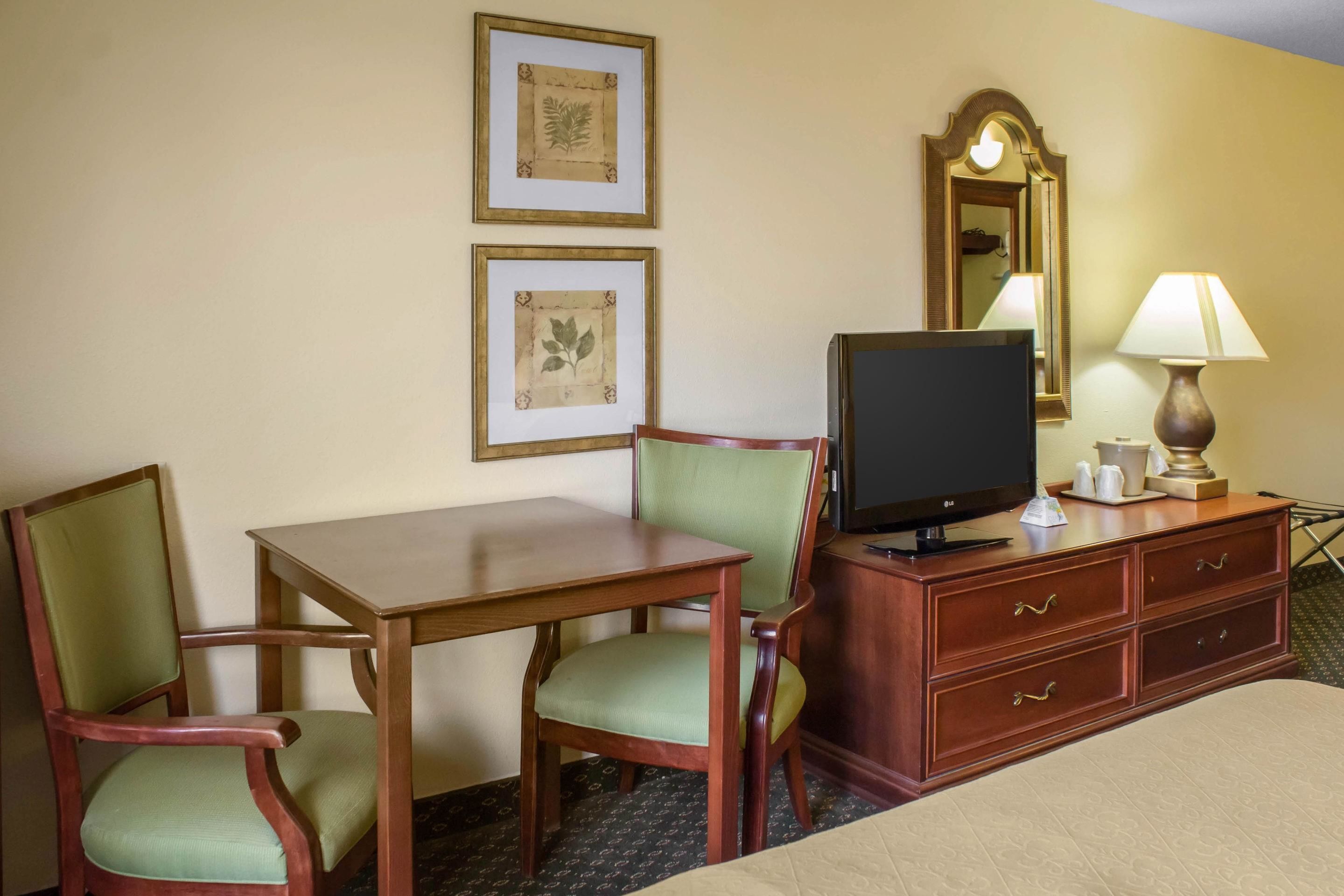 A hotel room with a wooden desk, two green chairs, a television, a mirror, a lamp, and a bed.