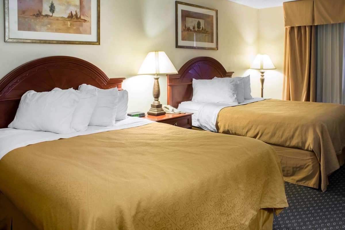 A hotel room with two beds, each with a wooden headboard, white bedding, and a brown throw blanket. There are two bedside tables with lamps, and the walls are decorated with framed artwork.