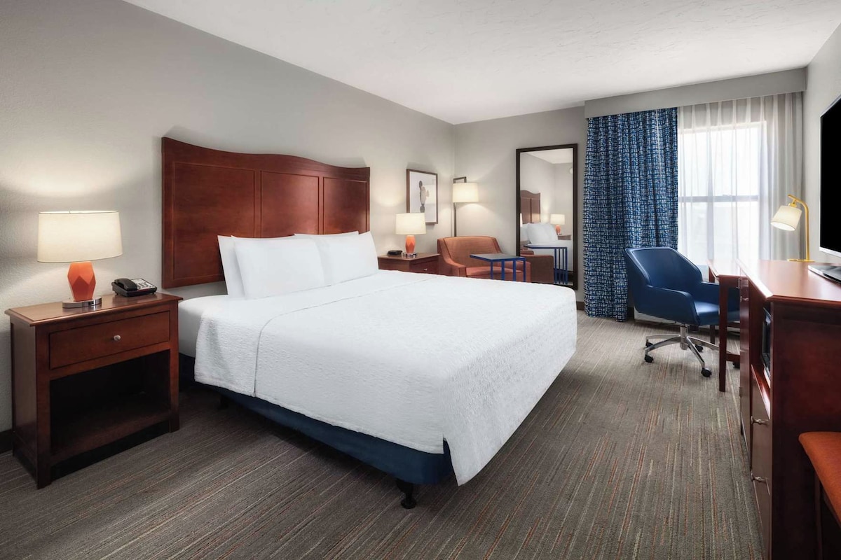 A hotel room with a large bed, bedside tables with lamps, a desk with a chair, a mirror, and a window with curtains.