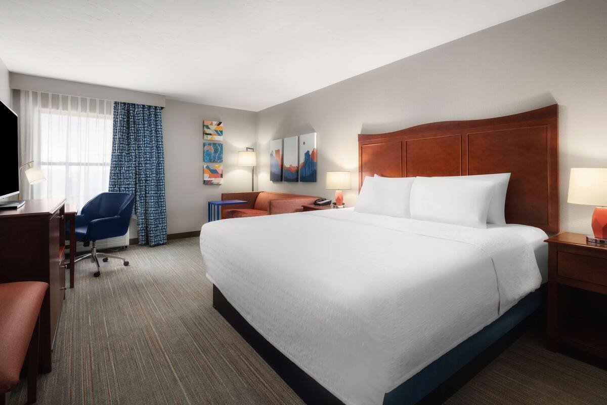 A hotel room with a large bed, a desk with a chair, a TV, and artwork on the walls.
