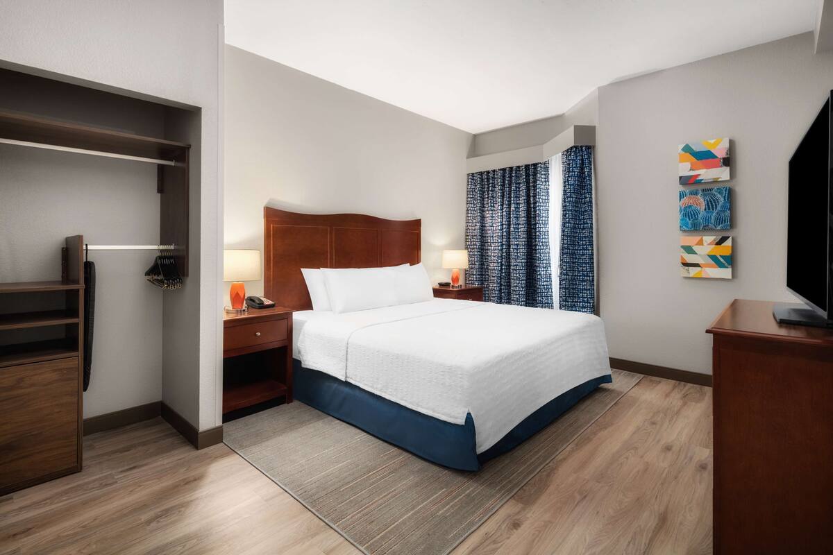A hotel room with a bed, nightstands, a desk, a wardrobe, and artwork on the wall.