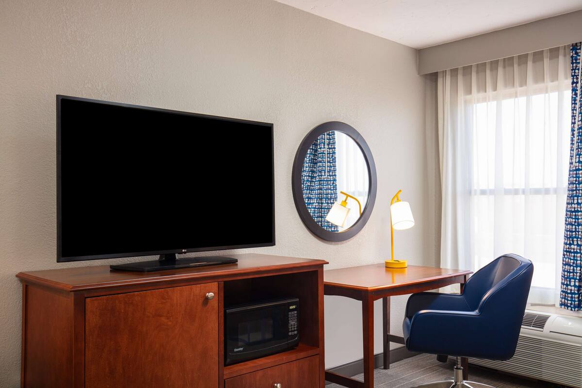 A hotel room with a flat-screen TV on a wooden cabinet, a desk with a chair, a round mirror, and a window with curtains.