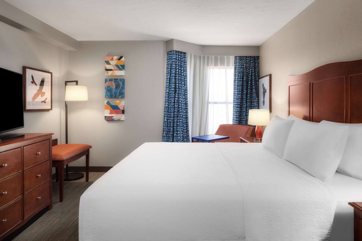 A hotel room with a large bed, a desk, a chair, a TV, and artwork on the walls.