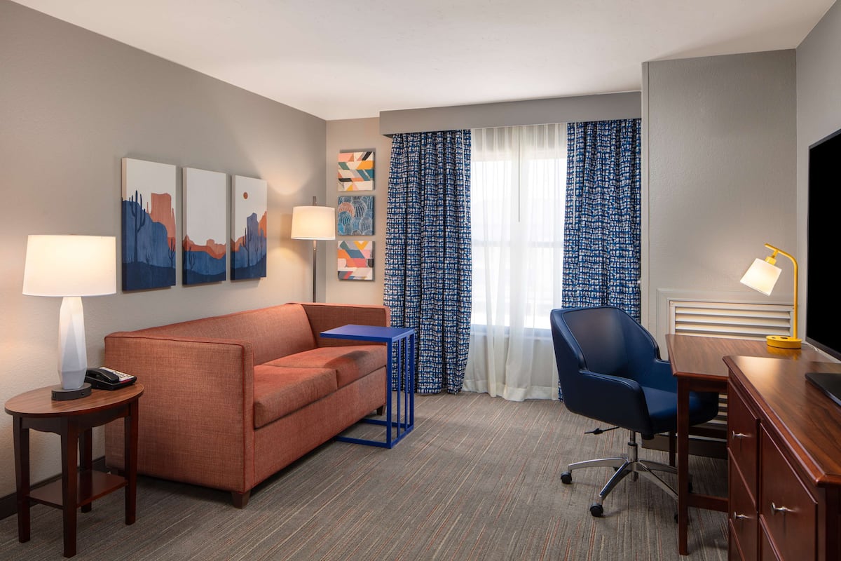 A hotel room with a sofa, a desk, a chair, a TV, and artwork on the walls.