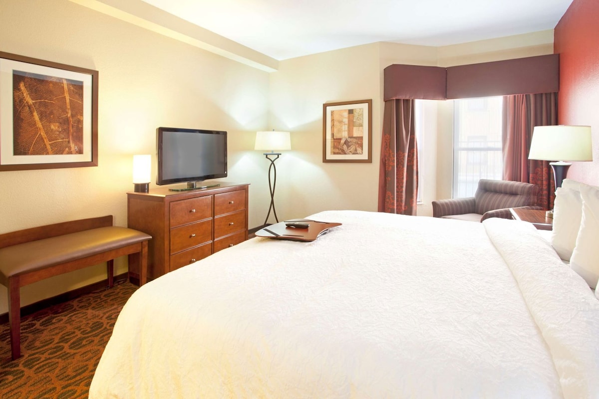 A hotel room with a large bed, a television, a desk with a lamp, a chair, and artwork on the walls.