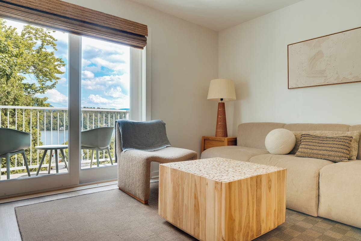 A modern living room with a sofa, a coffee table, and two chairs. There is a balcony with a view of trees and a railing.