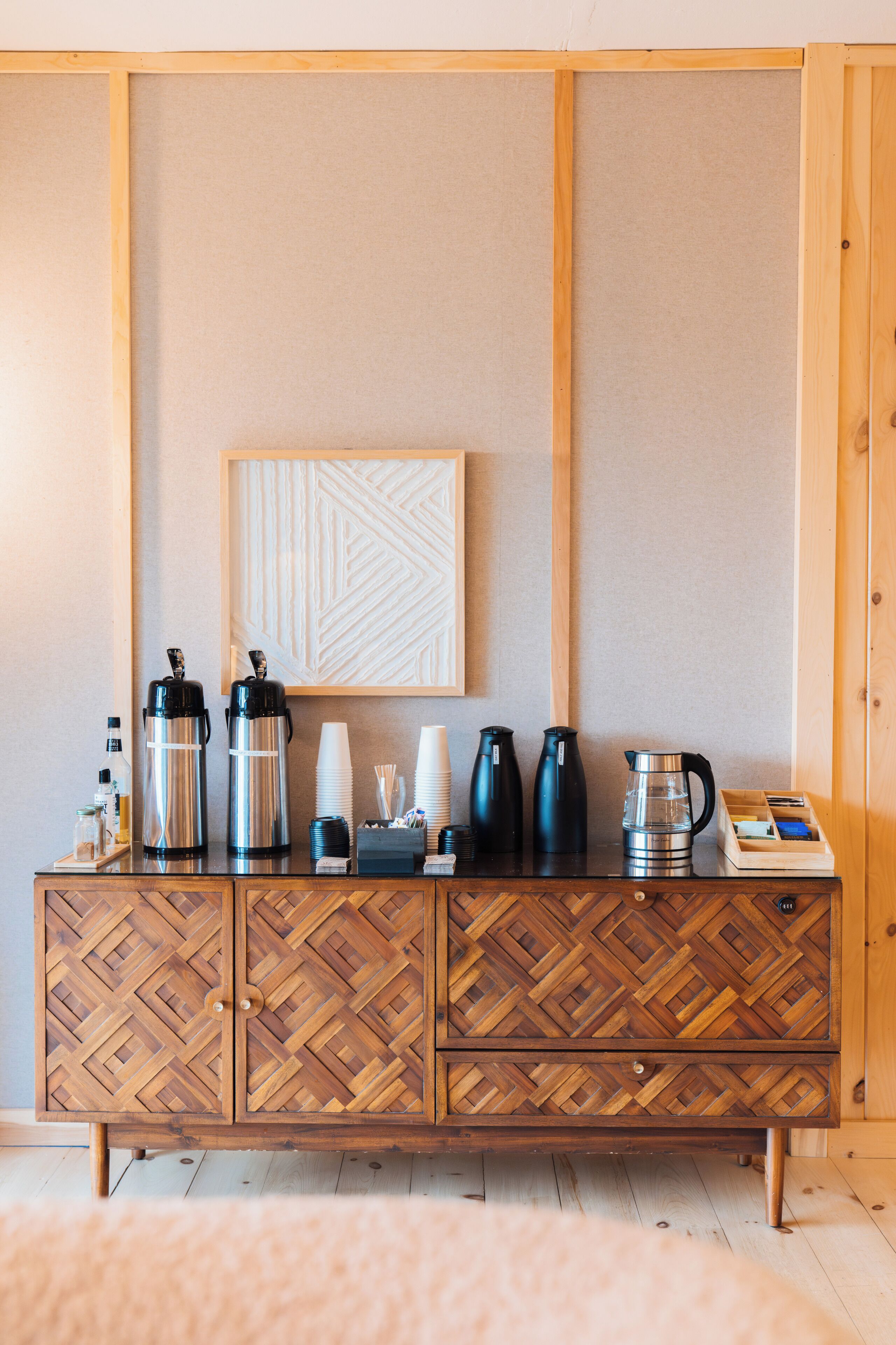 A wooden cabinet with a herringbone pattern, a coffee station with various coffee-making equipment, and a framed geometric artwork on the wall.