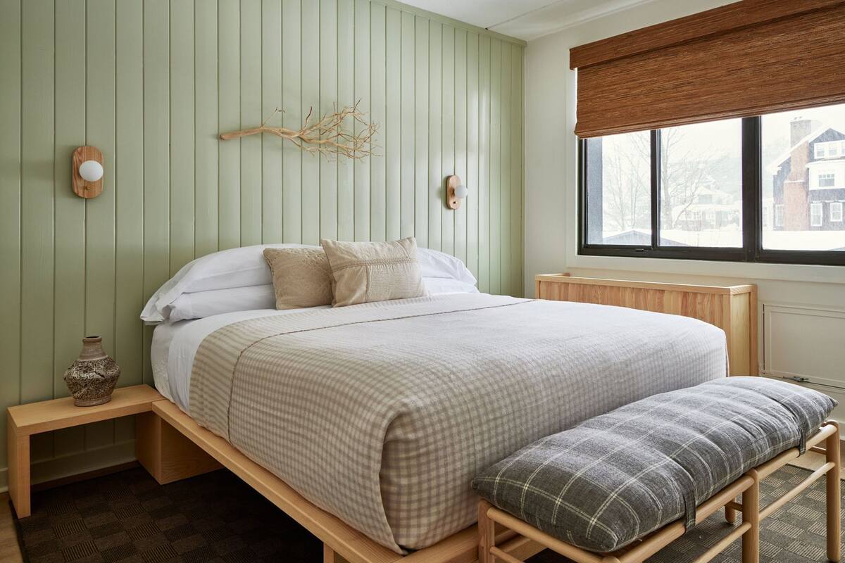 A bedroom with a bed, bedside tables, a window, and a view of buildings outside.