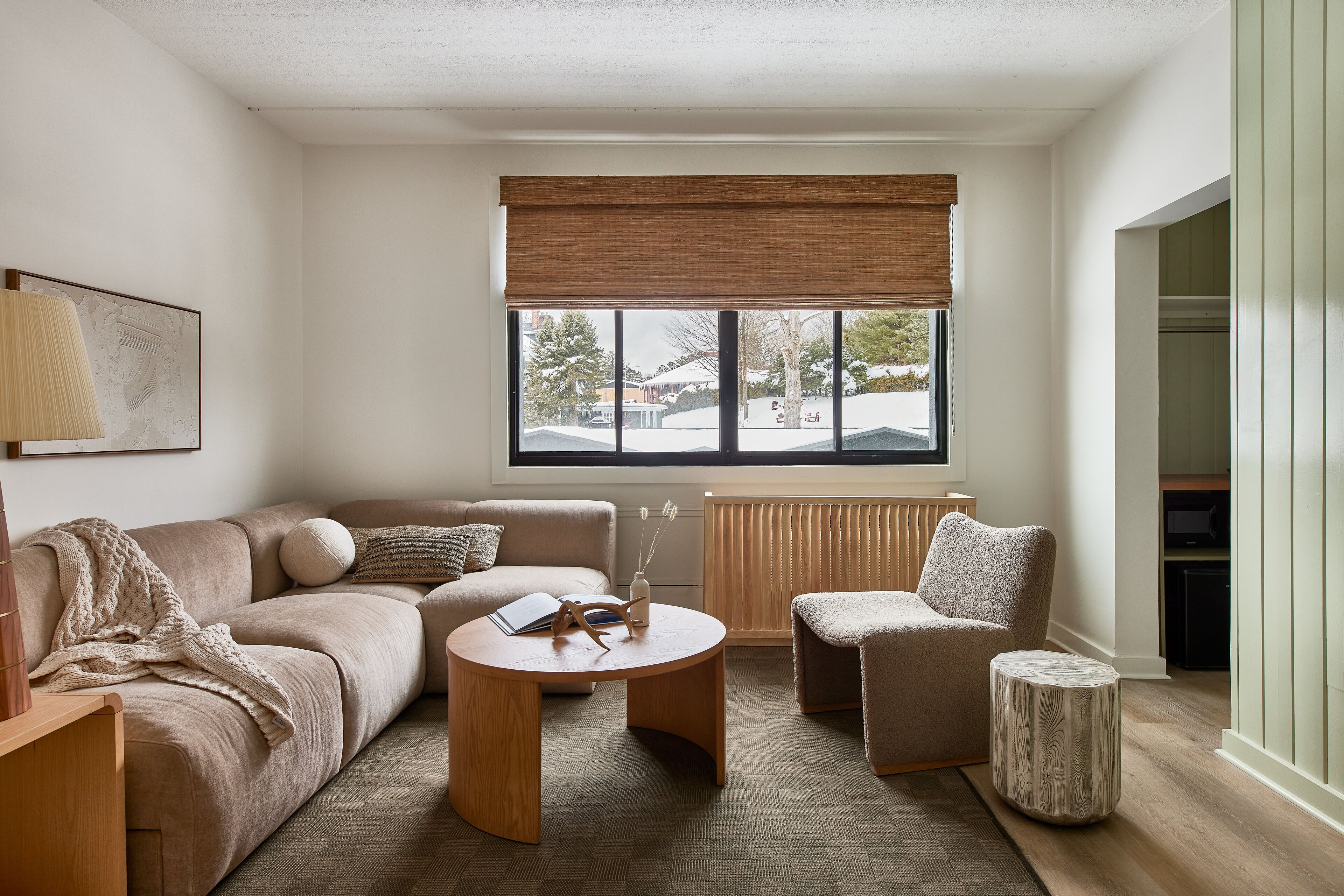 A modern living room with a beige sofa, a round coffee table, and a small side table. A large window with a wooden blind allows natural light to enter.