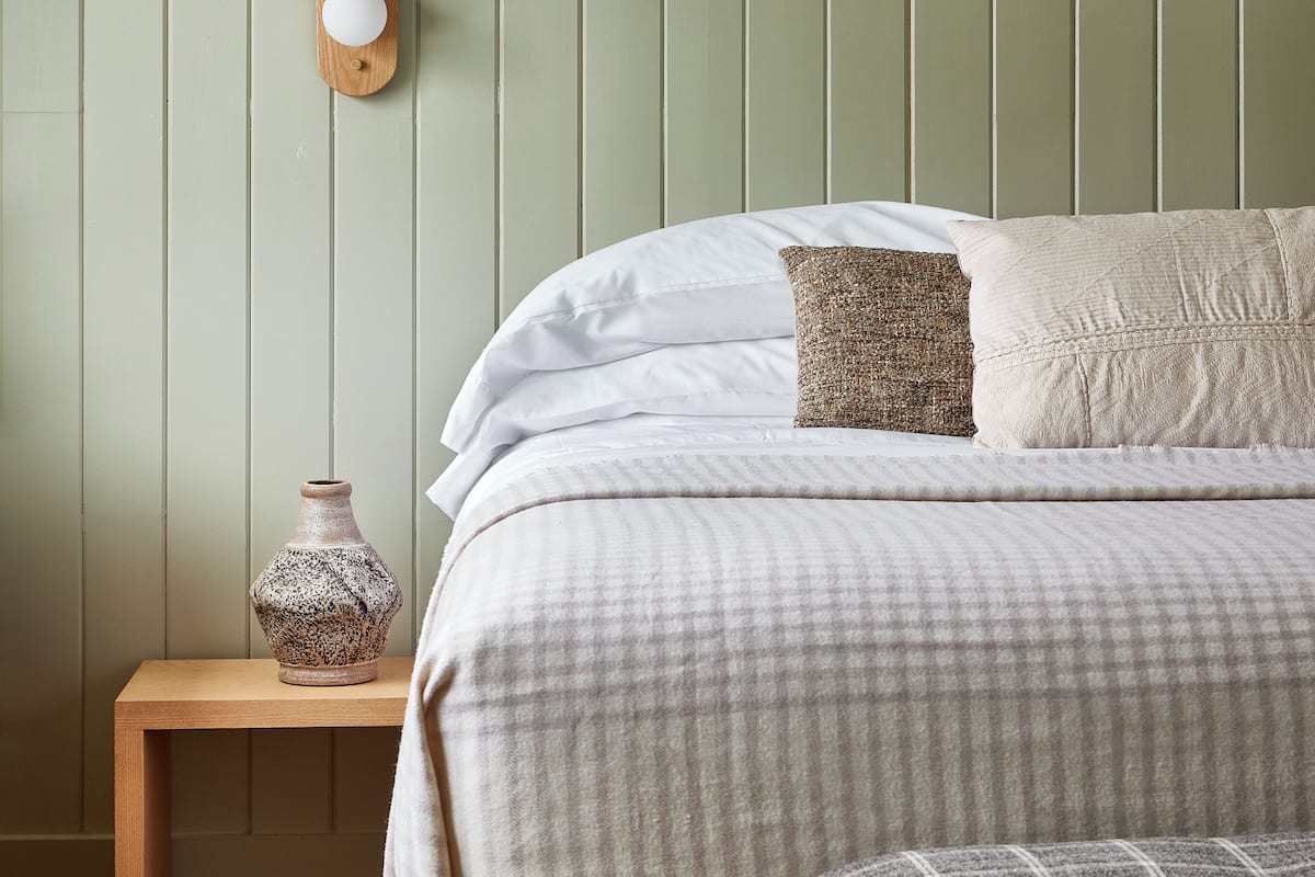 A neatly made bed with a checkered blanket, a bedside table with a ceramic vase, and a branch decoration on the wall.