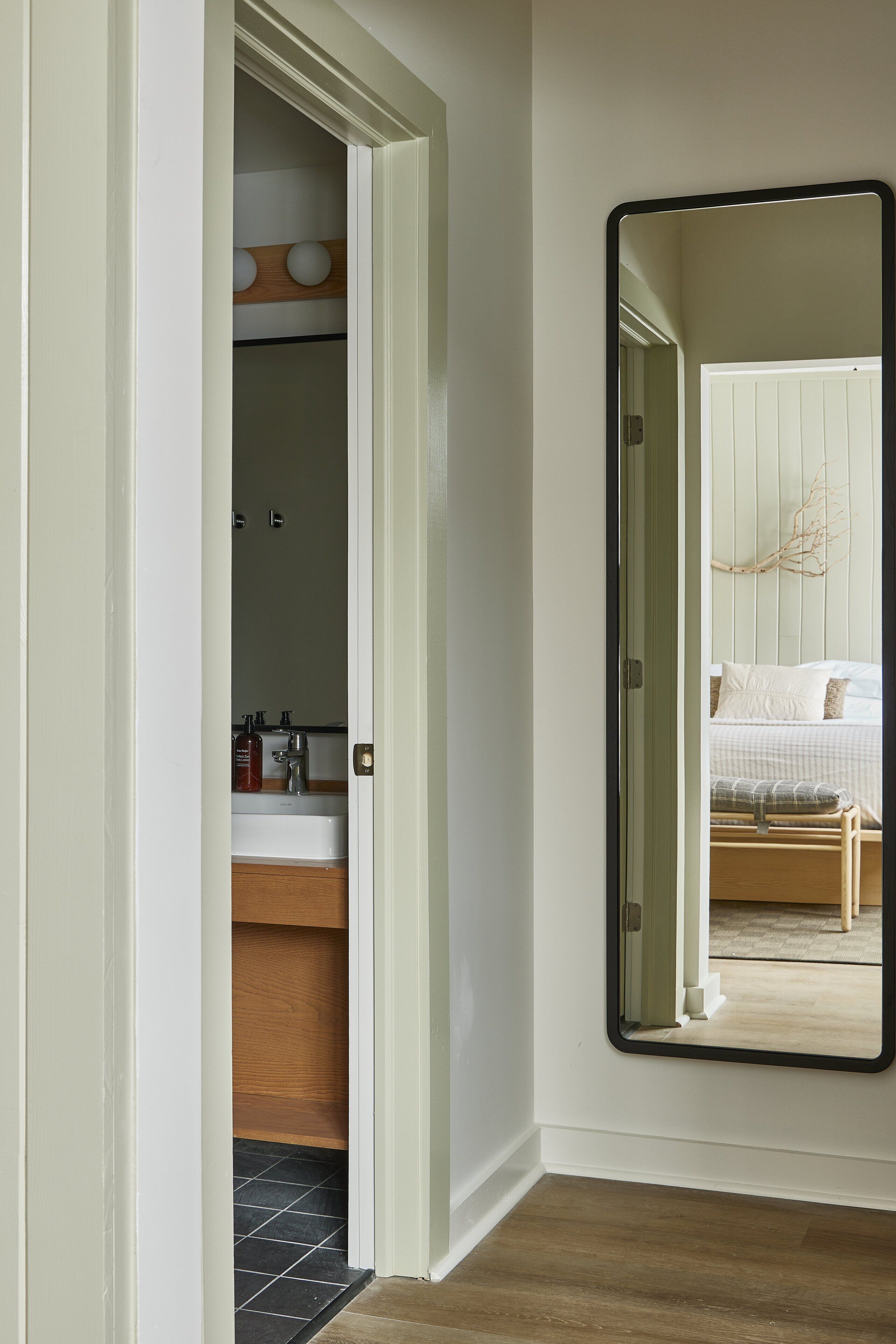 A hallway with a mirror reflecting a bedroom. The room has a bed, a nightstand, and a dresser.