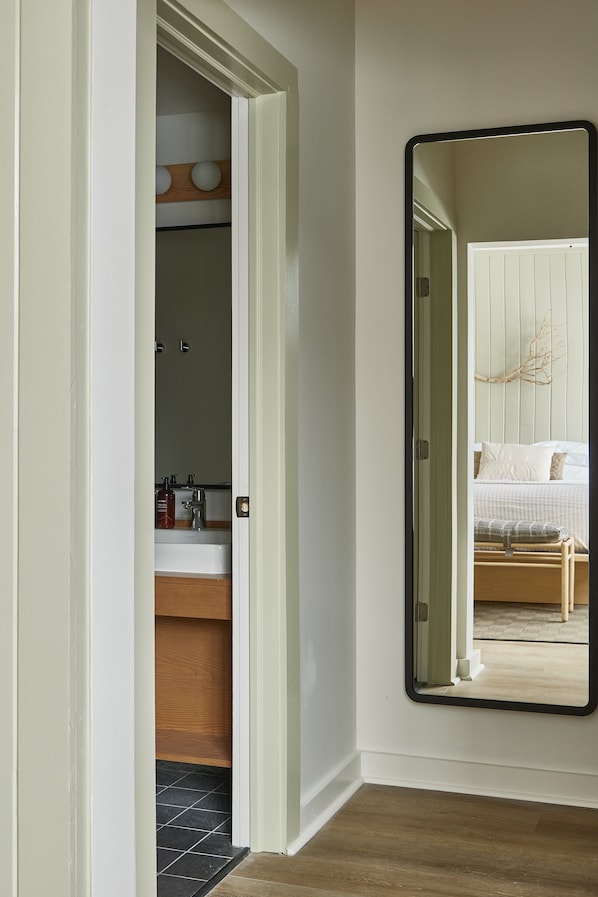 Un pasillo con un espejo que refleja un dormitorio. La habitación tiene una cama, una mesita de noche y un aparador.