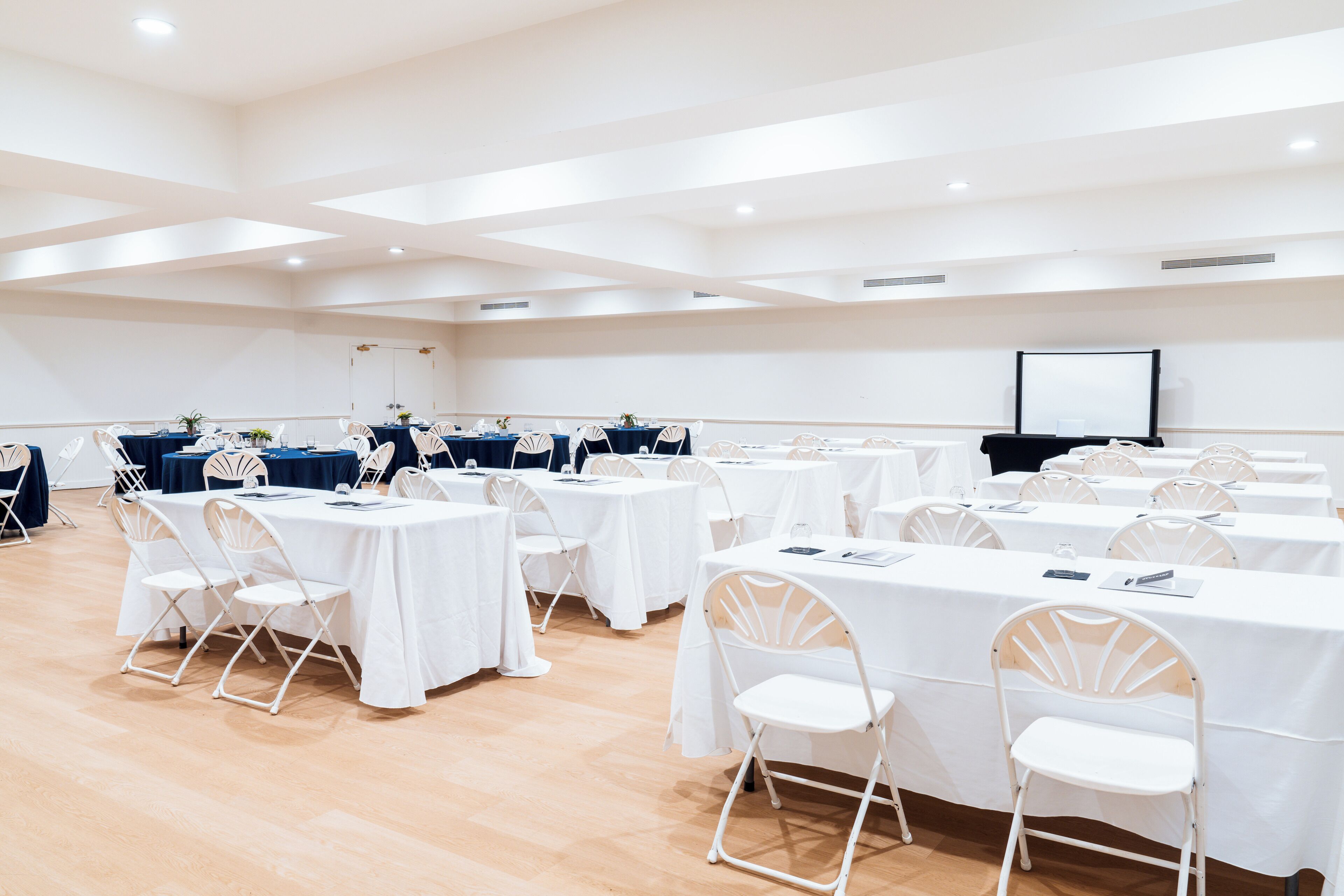 A conference room with multiple tables and chairs, a projector screen, and a wooden floor.