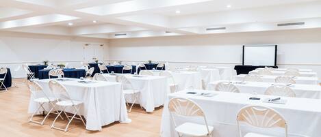 A conference room with multiple tables and chairs, a projector screen, and a wooden floor.