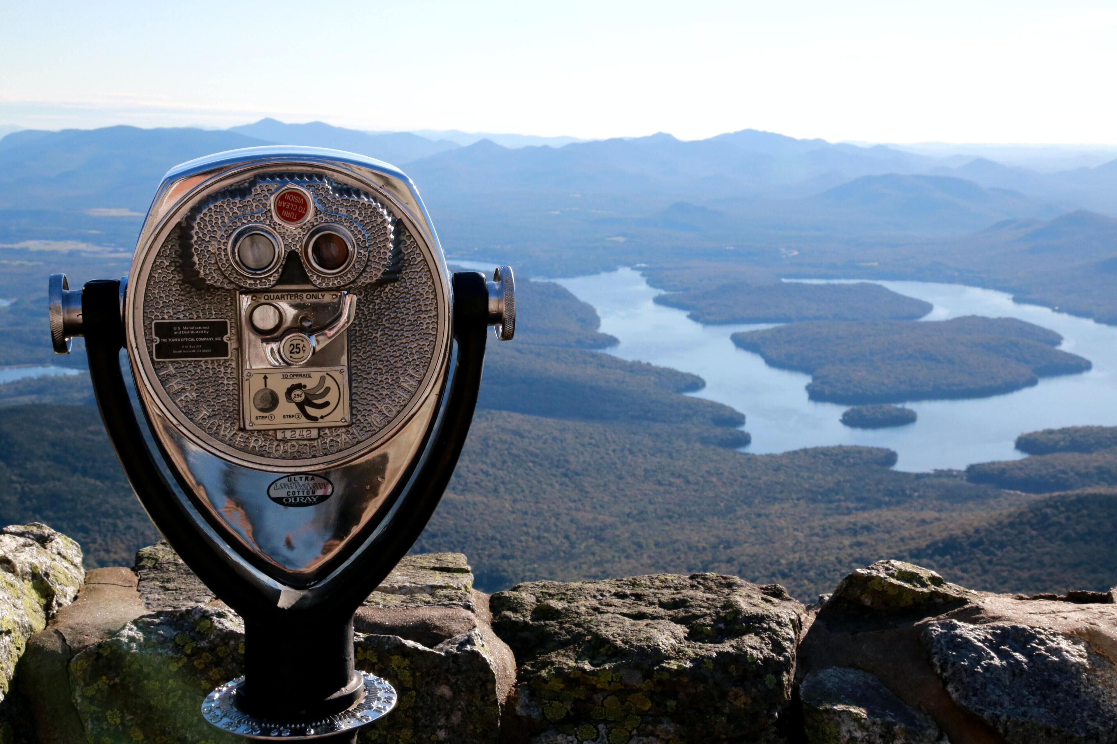 Ett gammalt teleskop med ett metallhölje och en svart bas, placerat på en klippavsats med utsikt över en sjö som omges av berg.