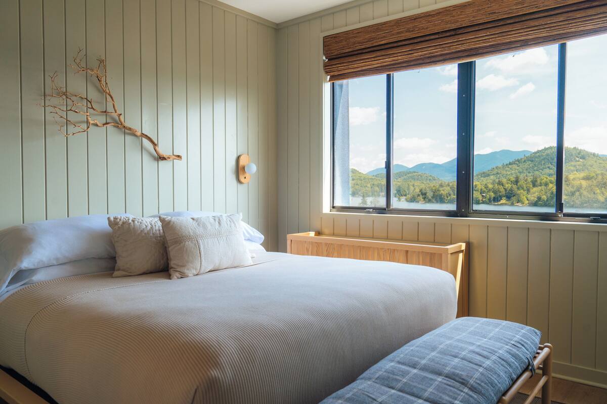 A bedroom with a large bed, a bench, a view of mountains through a window, and a wall-mounted branch decoration.