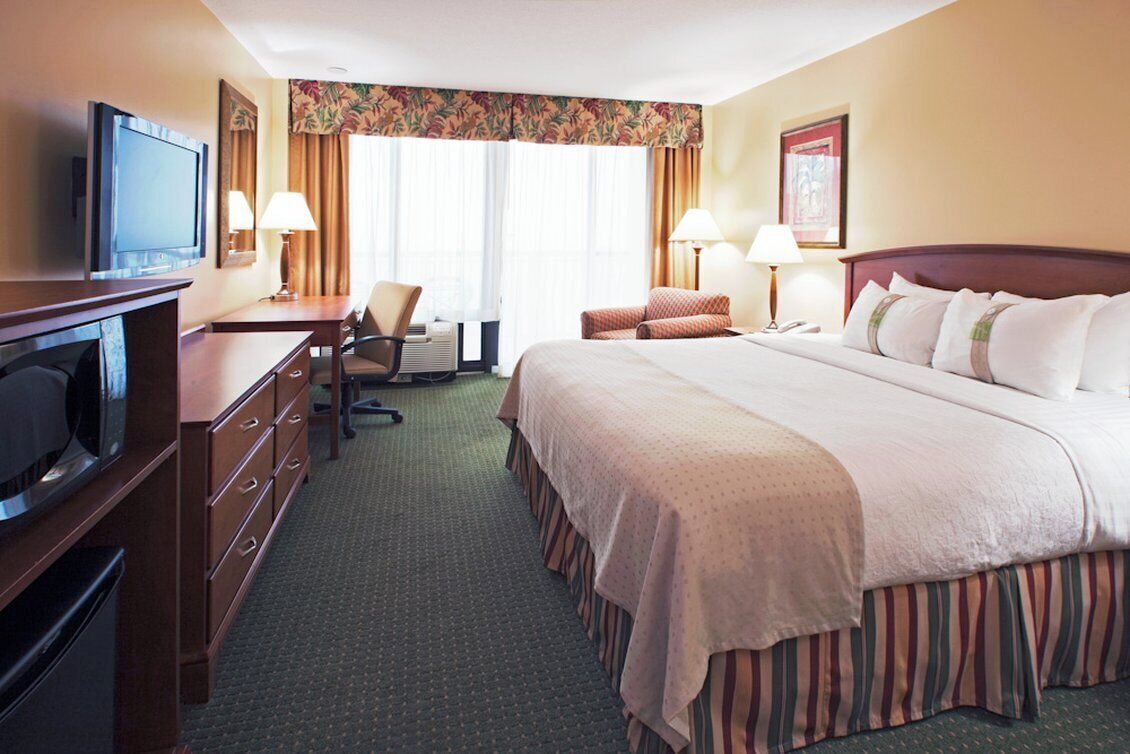 A hotel room with a bed, a desk with a television, a chair, and a window with curtains.