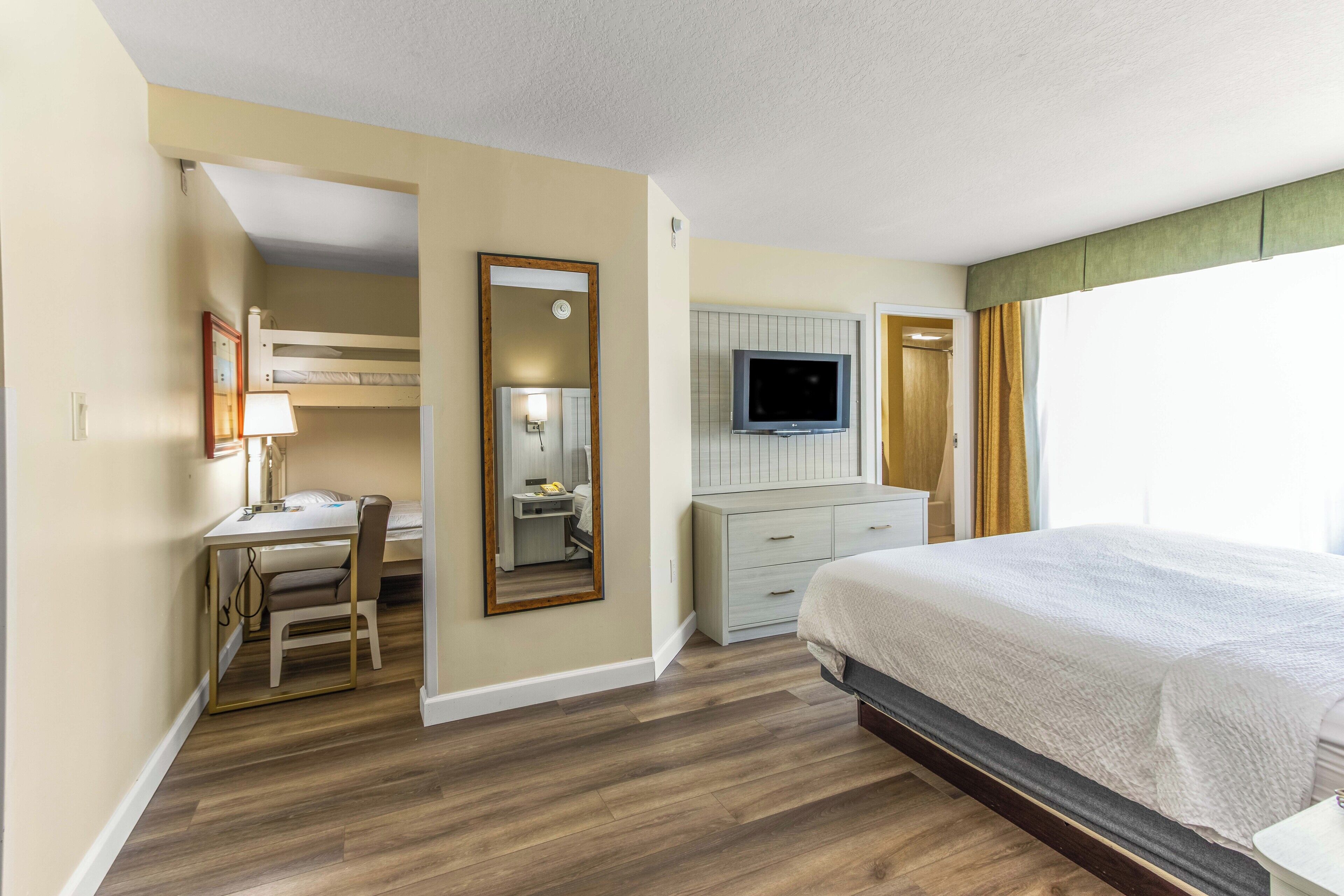 A hotel room with a bunk bed, a desk with a chair, a television, a dresser, and a large bed.