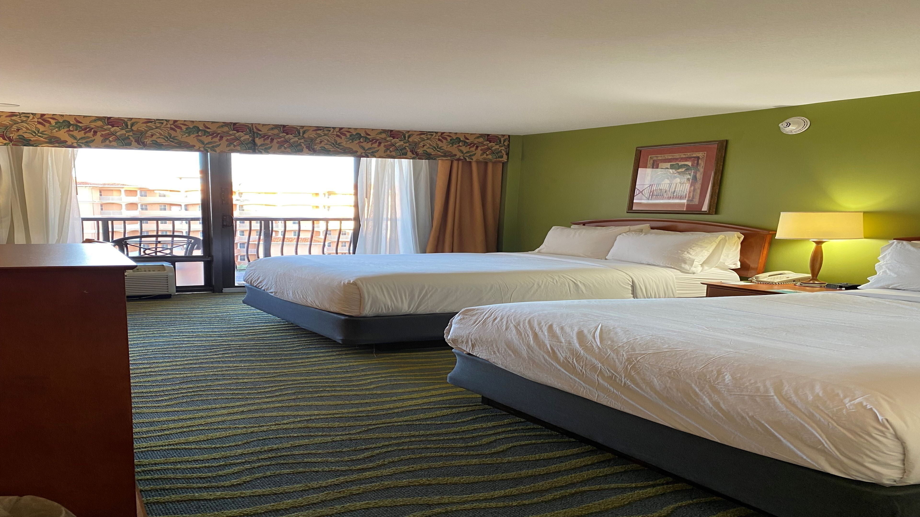 A hotel room with two beds, a desk, a chair, a lamp, and a view of the outside.