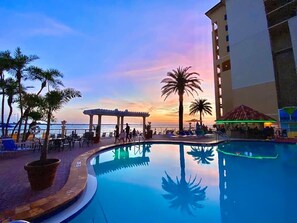 Un espace piscine offrant une vue sur le coucher de soleil, agrémenté de palmiers et bordé d’un bâtiment doté de balcons.