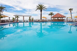 Une piscine d’un bleu éclatant, bordée de chaises longues et d’ombrelles, entourée de palmiers et agrémentée d’un kiosque.