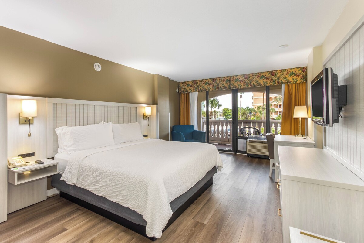 Habitación de hotel con una cama grande, mesitas de noche, un escritorio, una silla, un televisor y un balcón con vistas.
