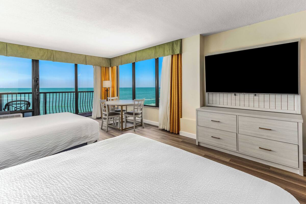 Ein Hotelzimmer mit zwei Betten, einem Esstisch, einem Fernseher und einem Balkon mit Meerblick.