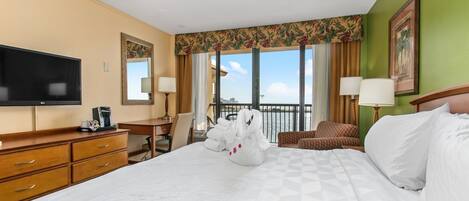 A hotel room with a large bed, a desk, a television, and a view of the sea.