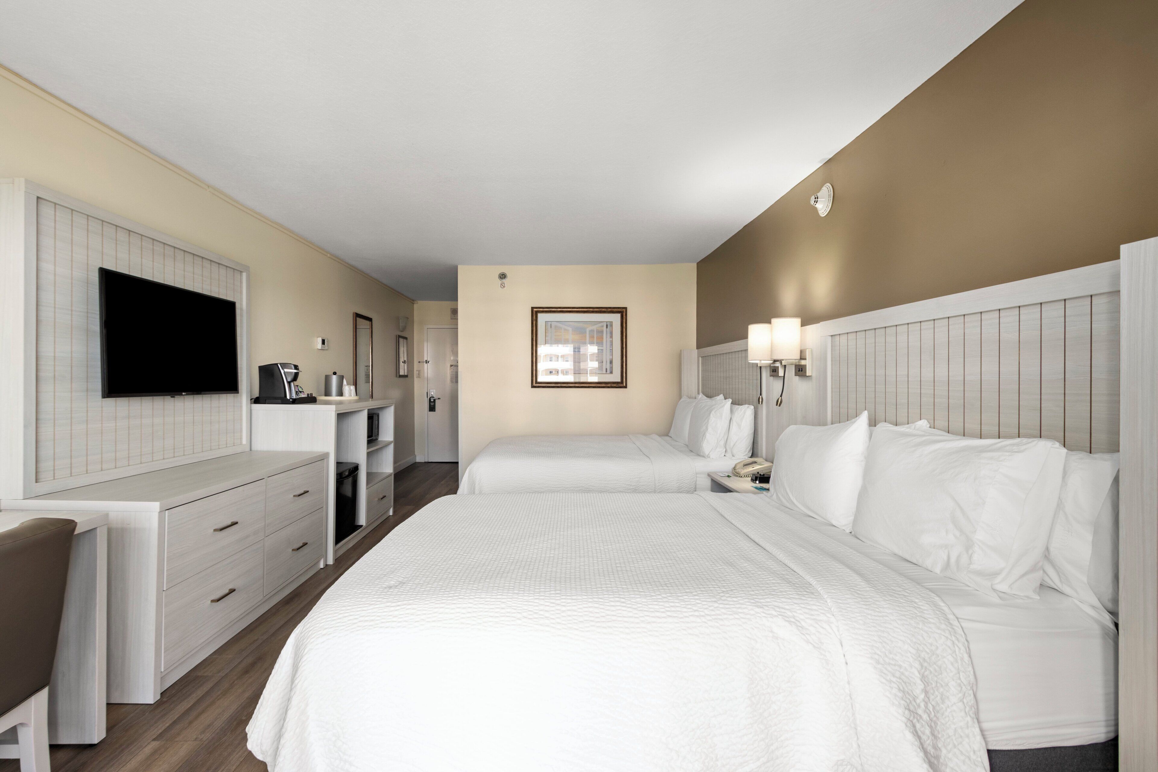 A hotel room with two beds, a desk with a flat-screen TV, a coffee machine, and a framed picture on the wall.