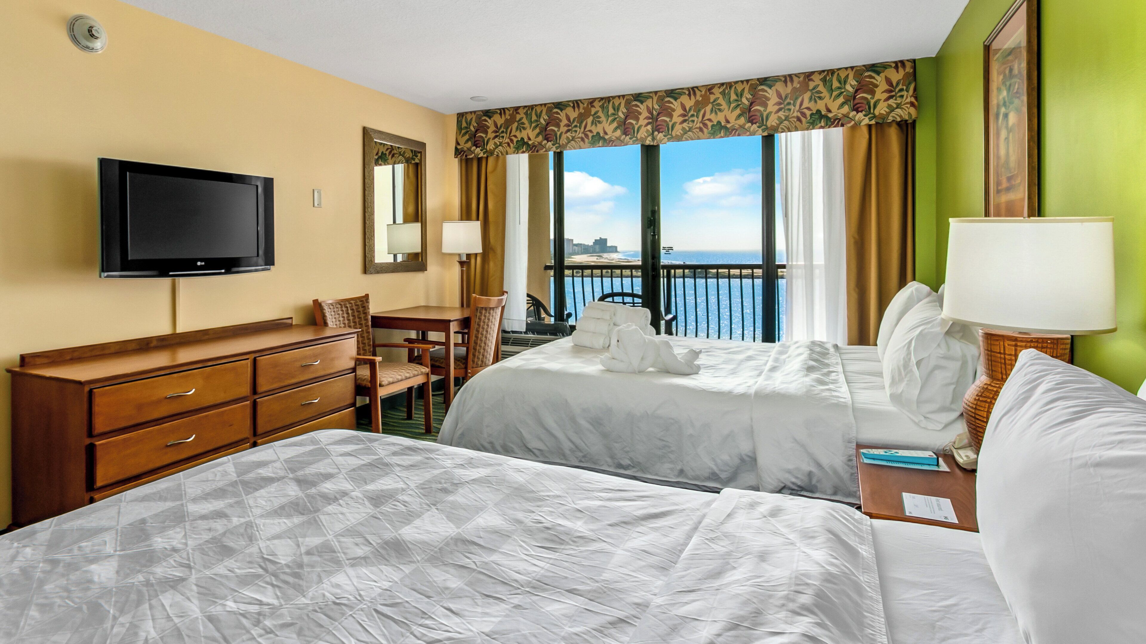 A hotel room with two beds, a TV, a desk, and a view of the ocean.