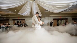 Recepção de casamento com um casal dançando em meio a uma nuvem de fumaça, cercado por convidados sentados em mesas.