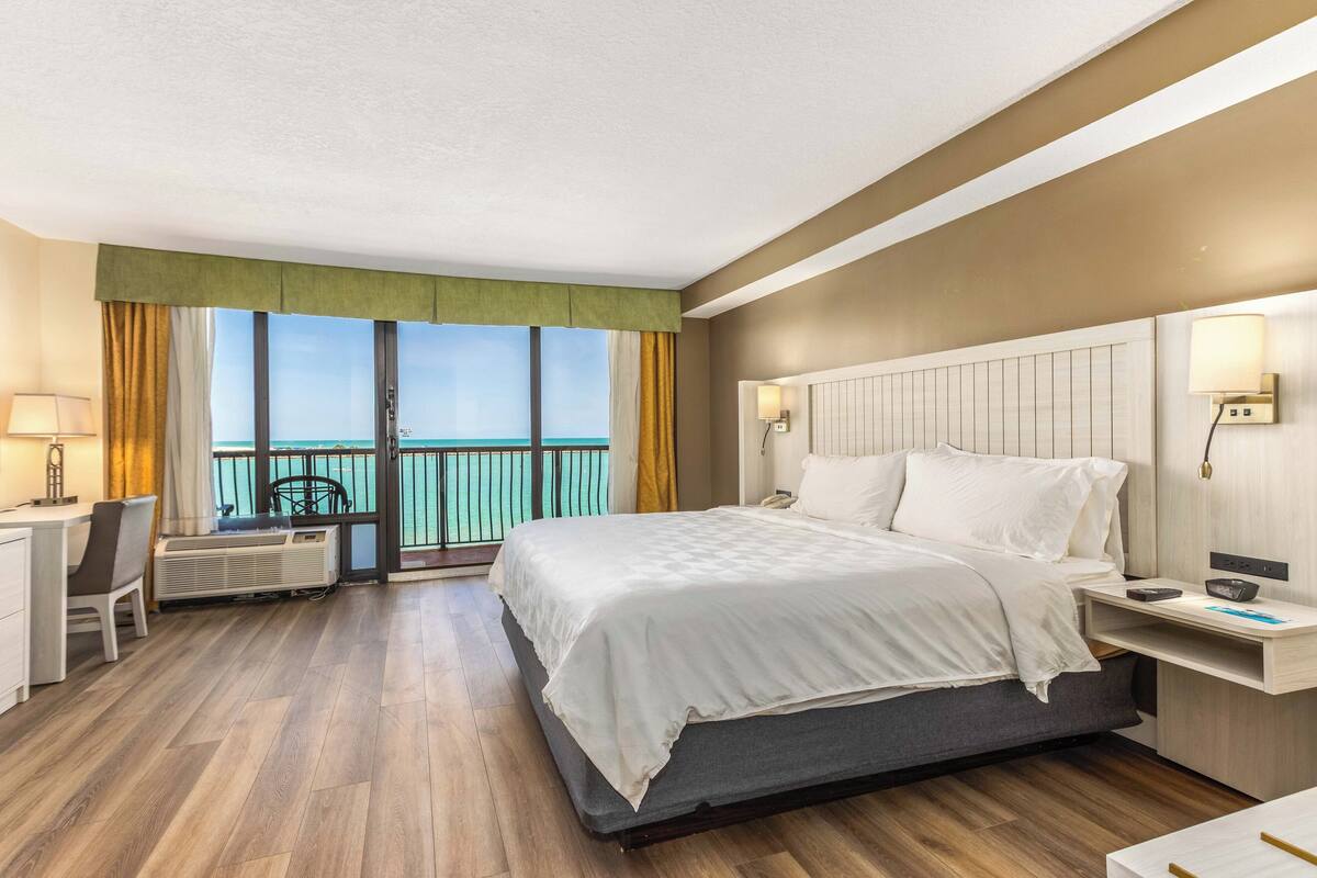 Ein Hotelzimmer mit Bett, Nachttisch, Lampe, Schreibtisch, Stuhl und Balkonblick.