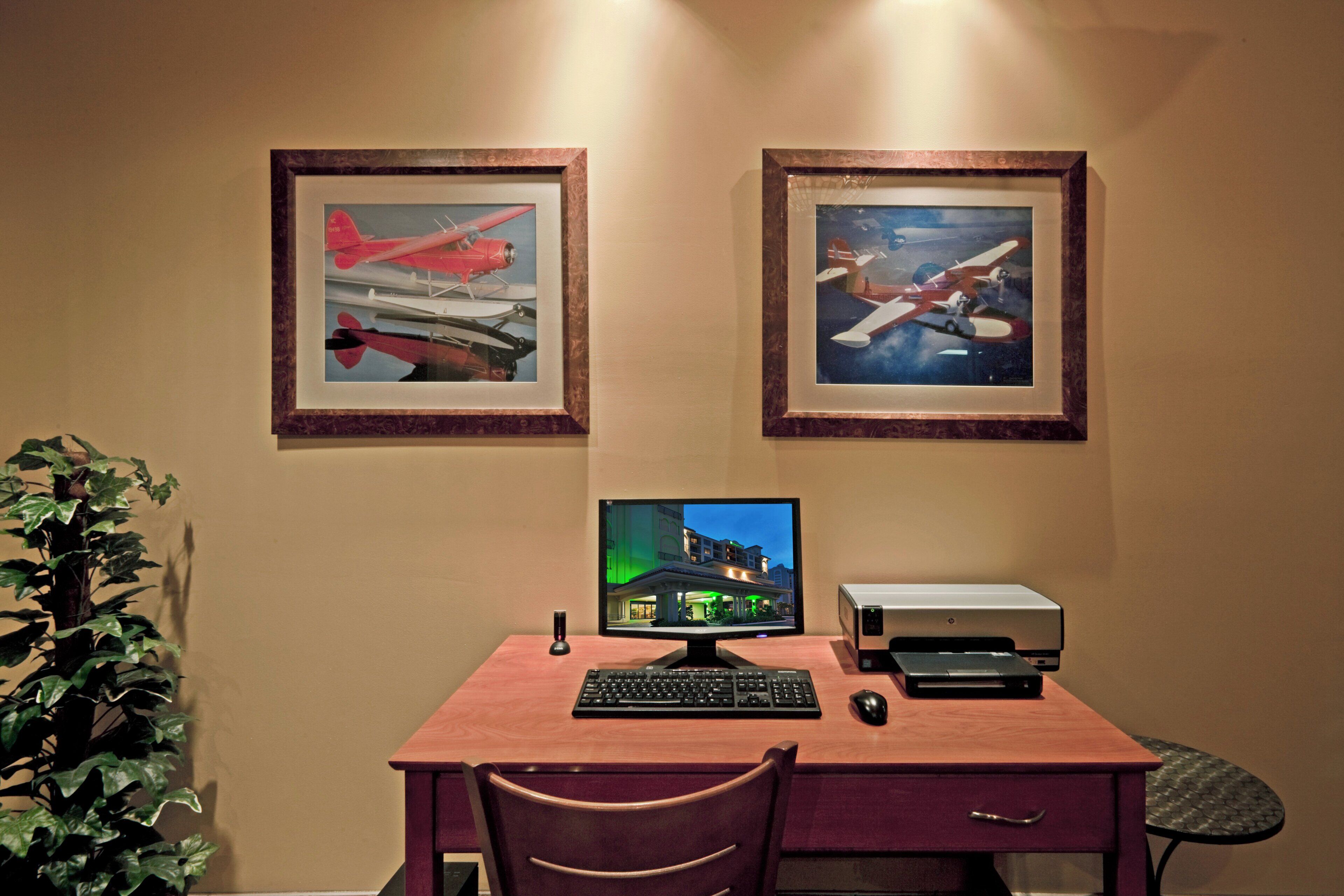 A desk with a computer, keyboard, mouse, and printer. There are two framed pictures of airplanes on the wall.