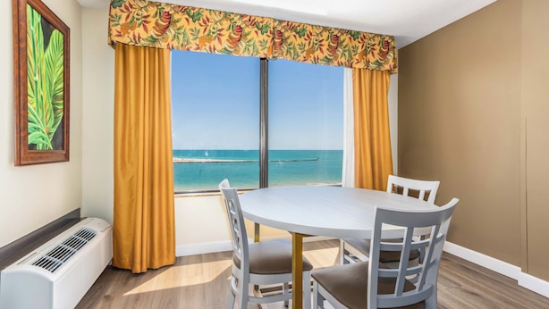 Uma área de refeições com mesa e cadeiras, uma janela grande com cortinas, um ar condicionado fixado na parede e um quadro emoldurado.