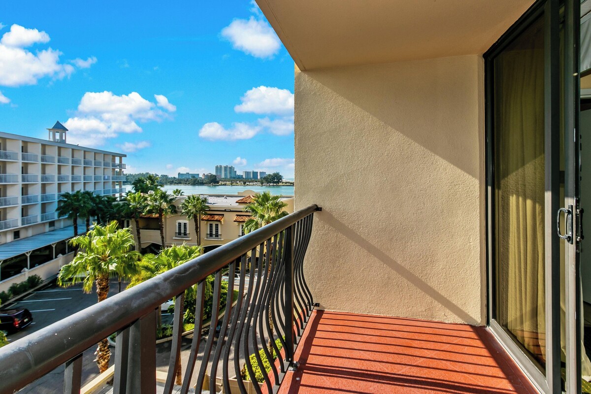 Ein Balkon mit rotem Boden, der auf ein Gewässer und eine Stadtansicht mit Gebäuden und Palmen blickt.