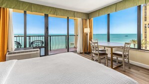 Une chambre d’hôtel avec un grand lit, une table à manger avec des chaises, un balcon avec vue sur la mer et une fenêtre avec des rideaux.