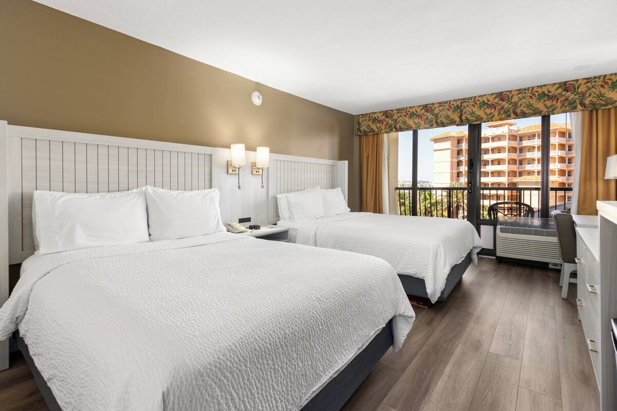 Ein Hotelzimmer mit zwei Betten, einem Schreibtisch und Blick auf ein Gebäude durch große Fenster.