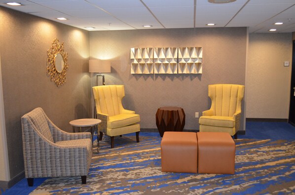 Un hall de hotel con dos sillones amarillos, un sillón de cuadros, una mesa redonda y un taburete de madera.