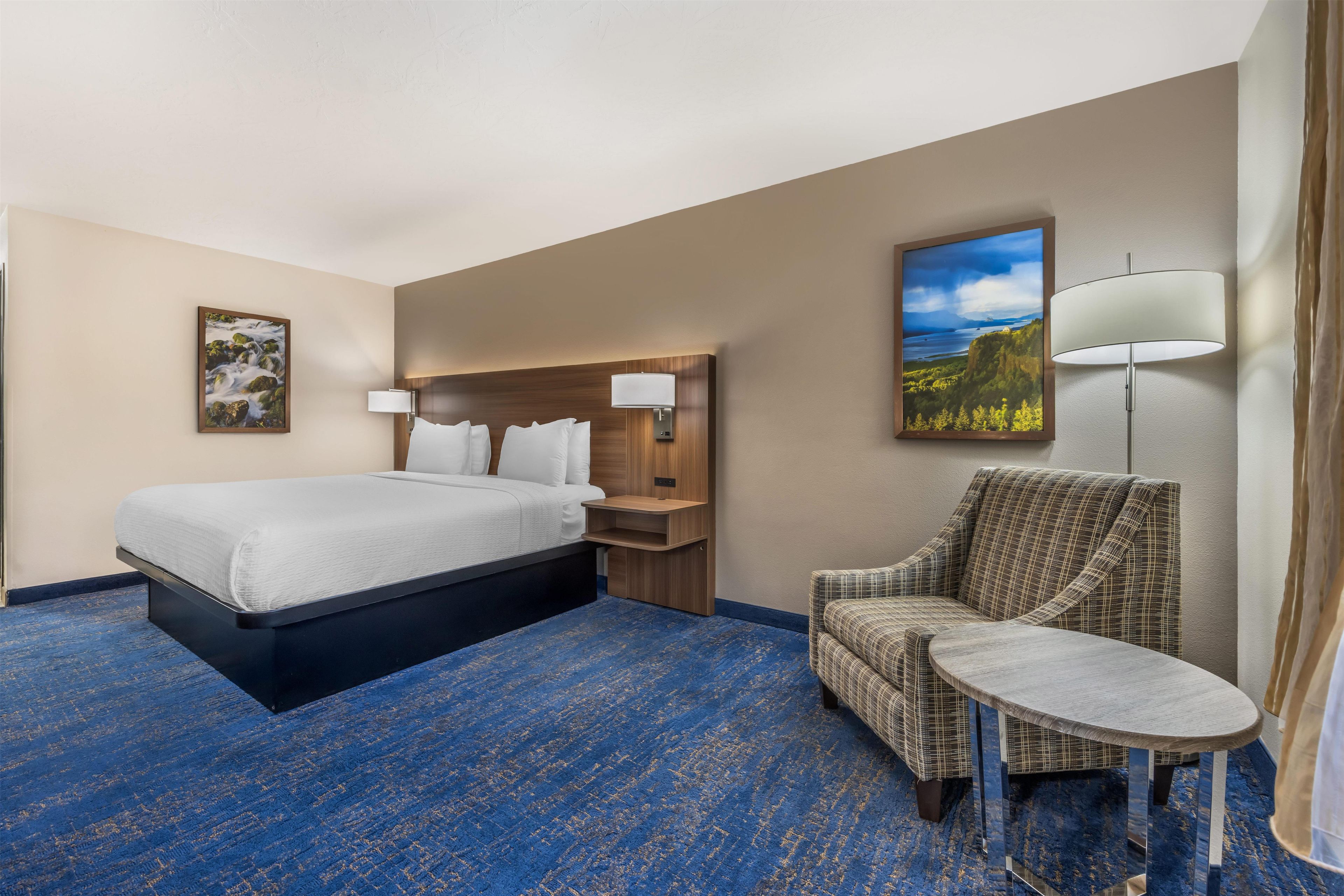 A hotel room with a bed, a chair, a small table, a nightstand, a lamp, and a painting on the wall.