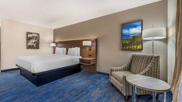A hotel room with a bed, a chair, a small table, a nightstand, a lamp, and a painting on the wall.