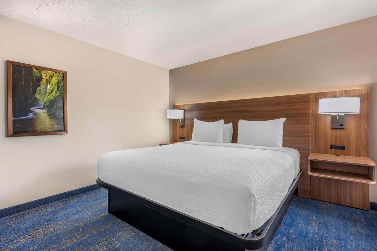 A hotel room with a large bed, two bedside tables, a nightstand, a wall-mounted lamp, and a painting of a natural scene.